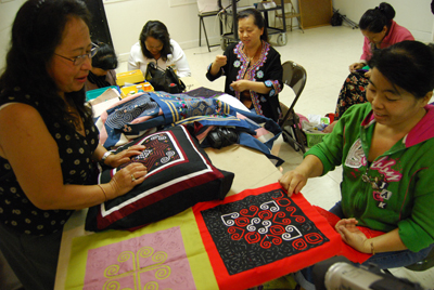Xong Lee is one of a group of women led by Ying Yang in weekly sewing circle gatherings focused on paj ntaub at the Homeland Cultural Center in Long Beach.  Master artist Yang (left, standing) gives feedback to apprentice Lee in fine detailed stitching as they examine the latest section of in her paj ntaub-in-process (bordered in red), while other sewing circle participants work on their individual projects in the background.  The paj-ntaub Lee is working on has snail and leaf patterns which Lee describes as “more complicated, more difficult because it involves more folding and more colors.  Each edge you have to turn in and sew, fold in and sew."