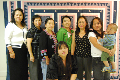 Some of the women of Homeland Cultural Center’s Hmong sewing circle led by master artist Yang stand in front of one of Yang’s full-sized paj ntaub pieces.  Left to right: Jenny Yang, Chou Yang, Mao Moua, Khou Lee (kneeling), apprentice Xong Lee, master artist Ying Yang, and Julia Yang holding Danny.